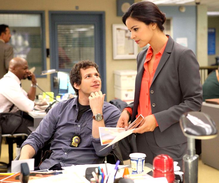 a woman standing next to a man sitting at a desk in an office setting with other people