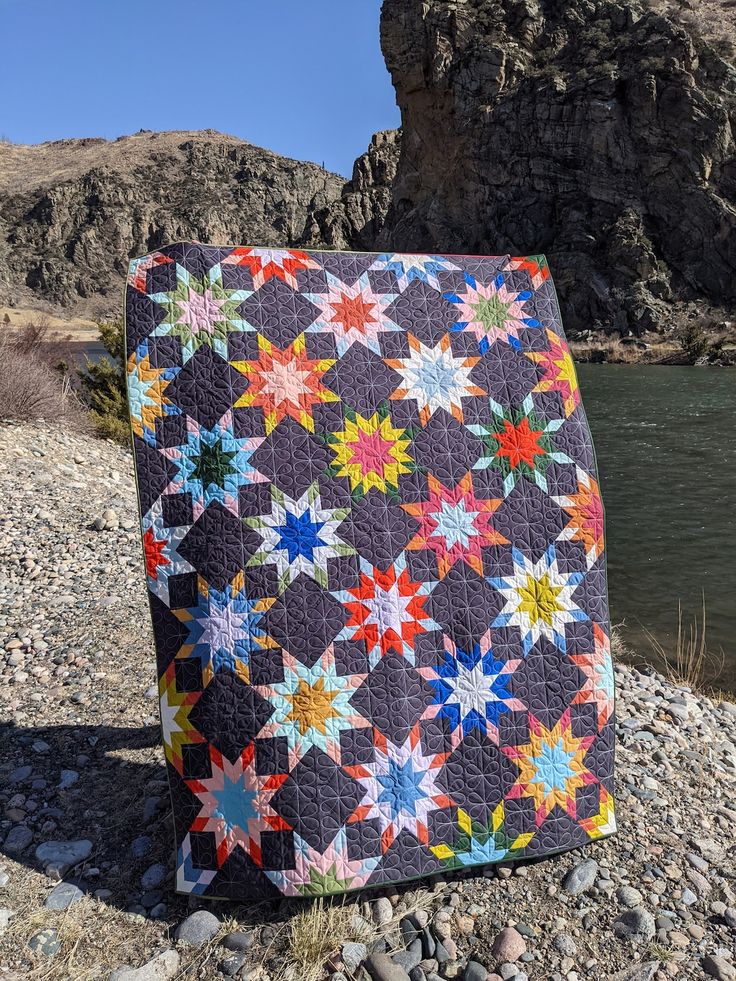 a colorful quilt sitting on top of a rocky beach next to a body of water