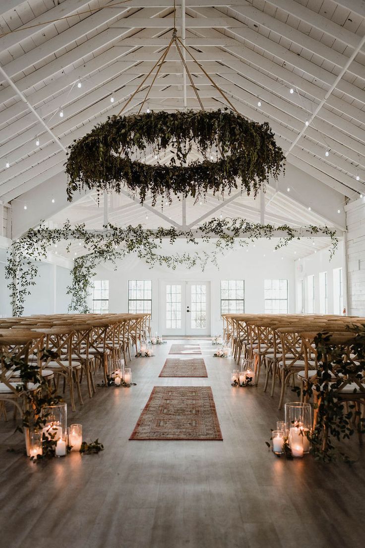 an indoor wedding venue with wooden chairs and greenery hanging from the ceiling, surrounded by candles