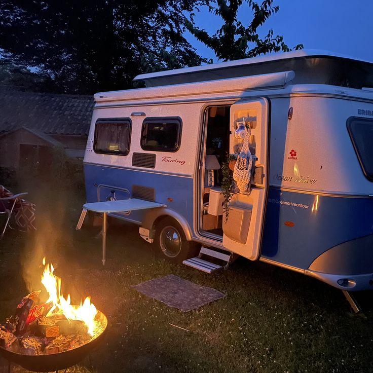 a camper parked next to a fire pit at night with the door open and it's lights on