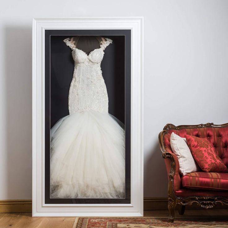 a white wedding dress hanging in a frame on the wall next to a red chair