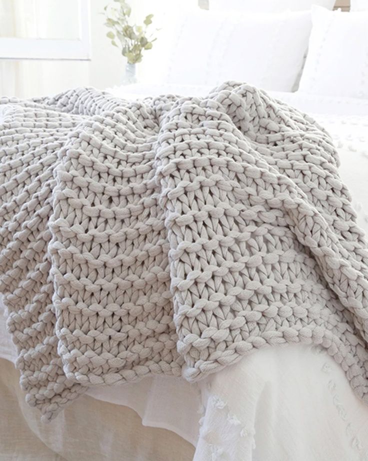 a white bed topped with a knitted blanket and pillows next to a vase filled with flowers