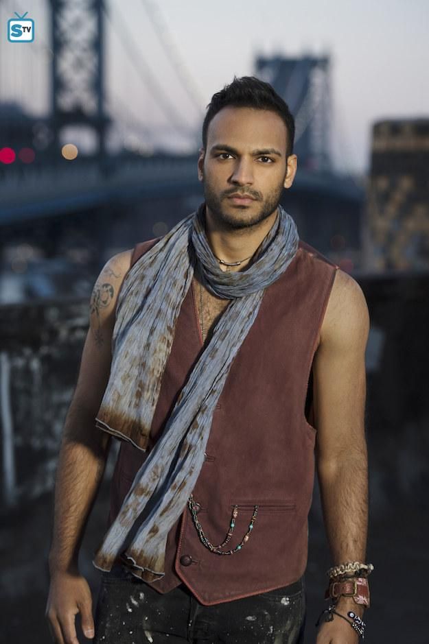 a man standing in front of a bridge wearing a brown vest and scarf with his hand on his hip