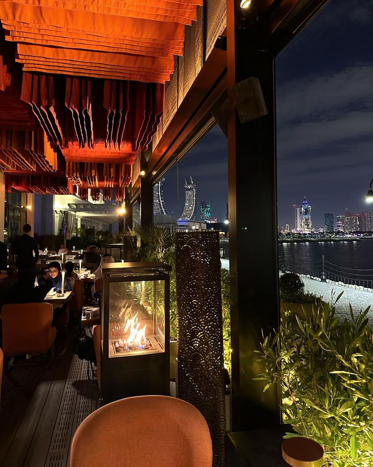 an outdoor restaurant with a fire place in front of the water and city lights at night