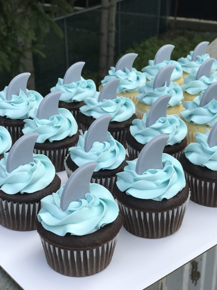 cupcakes with blue frosting and shark fin decorations are on a white table