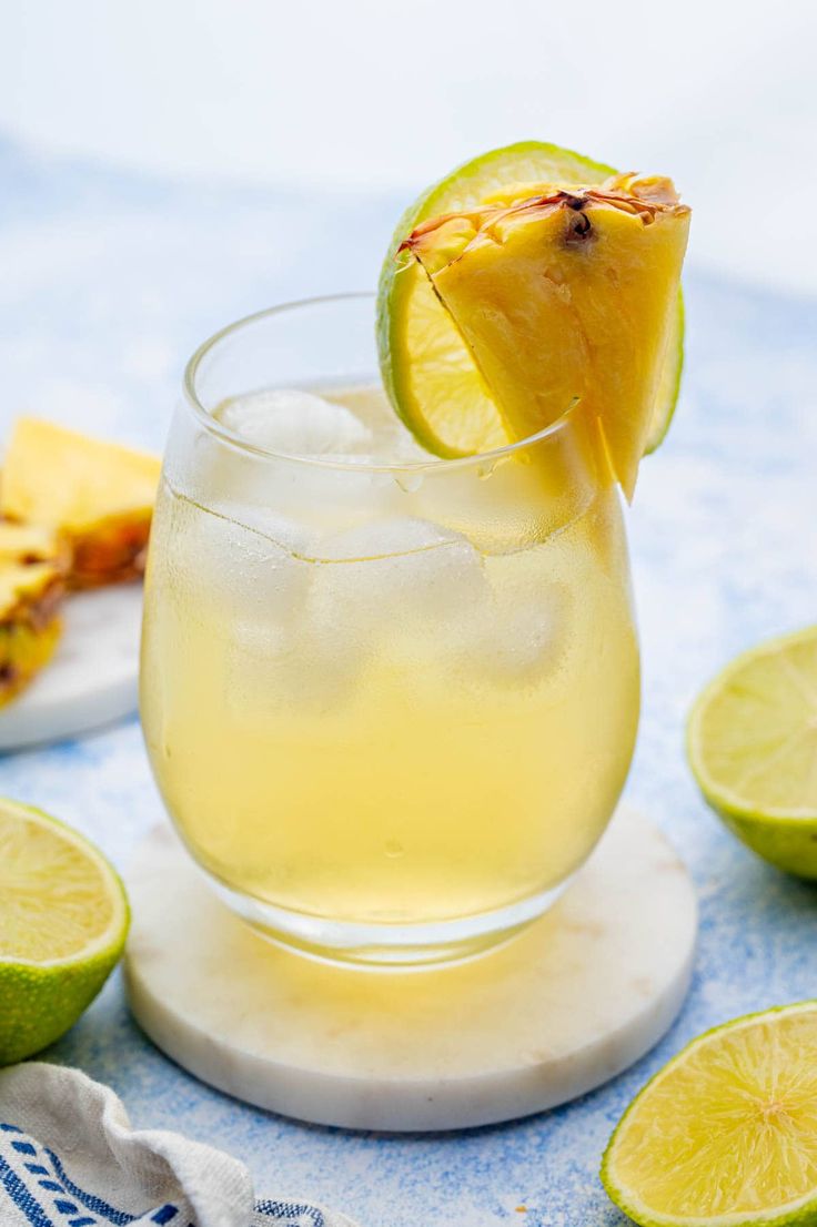 a close up of a drink in a glass on a table with limes and pineapple slices