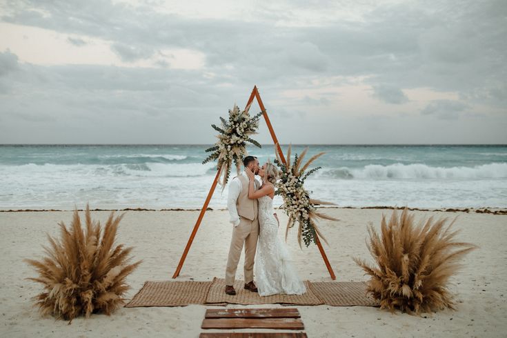 a bride and groom standing on the beach with their wedding arch decorated with greenery