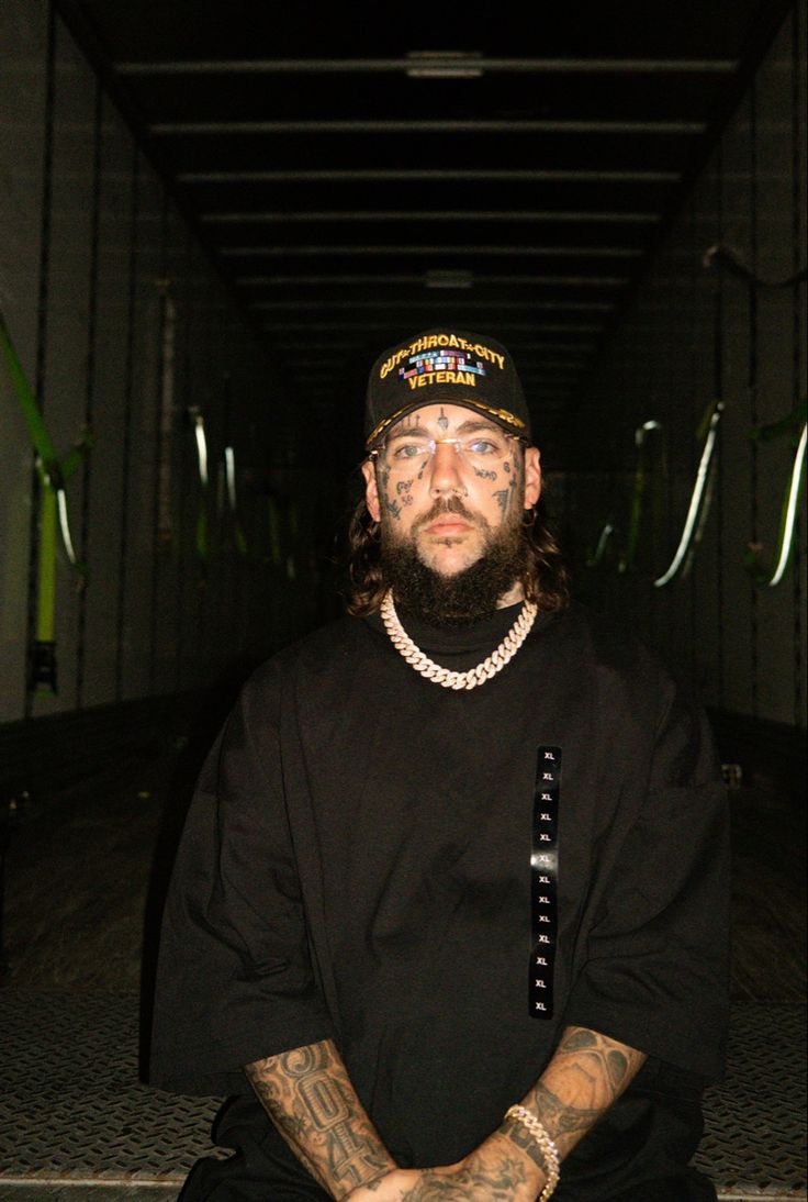 a man with tattoos and piercings sitting on the ground wearing a black t - shirt