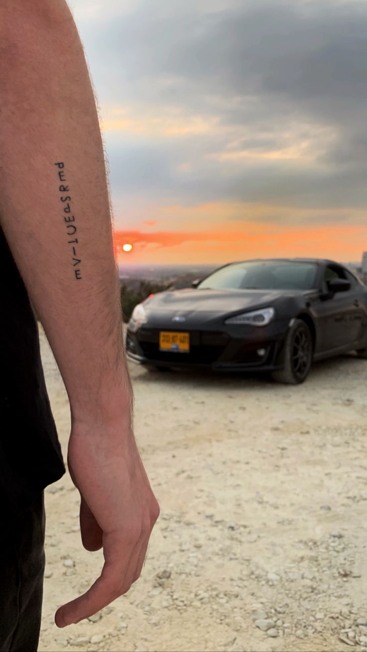 a man with a tattoo on his arm standing in front of a sports car at sunset