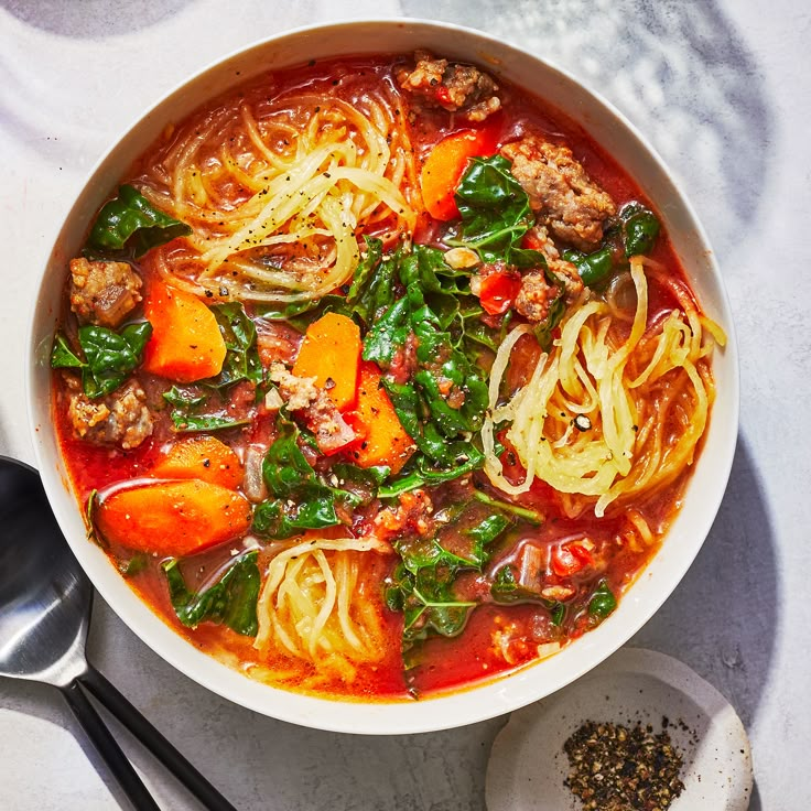 a bowl of soup with meat, noodles and spinach on the side next to two spoons