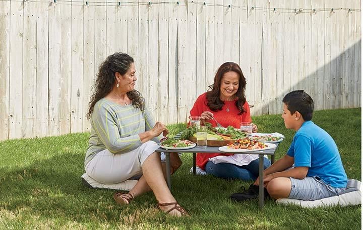 a woman and two children sitting at a table with food on it in the grass