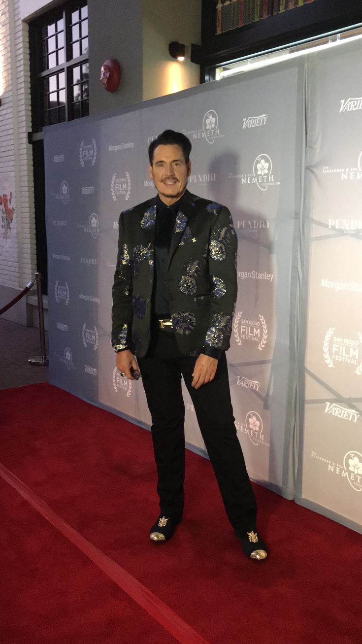 a man standing on top of a red carpet in front of a white wall wearing a suit and tie