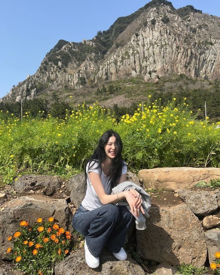 a woman sitting on top of a rock next to flowers and mountains in the background