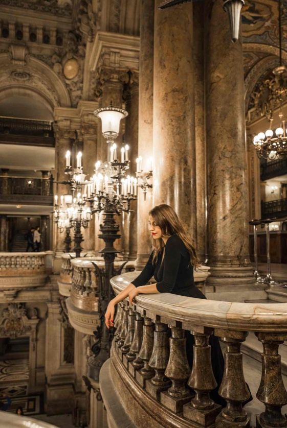 a woman is sitting on a balcony looking down at the chandelier and stairs