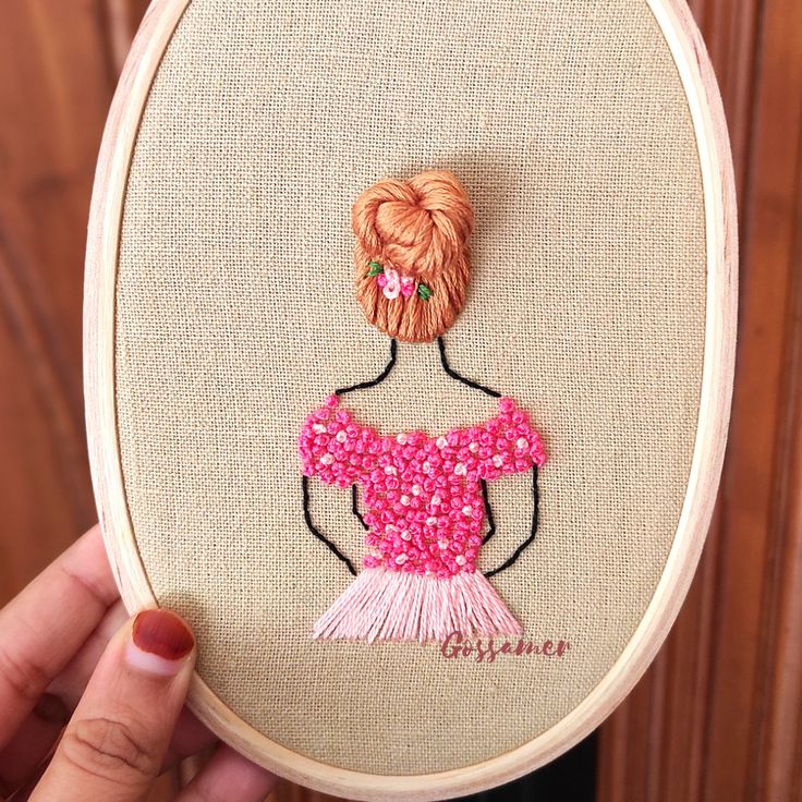 a woman's hand is holding up a cross - stitched picture with a pink dress on it