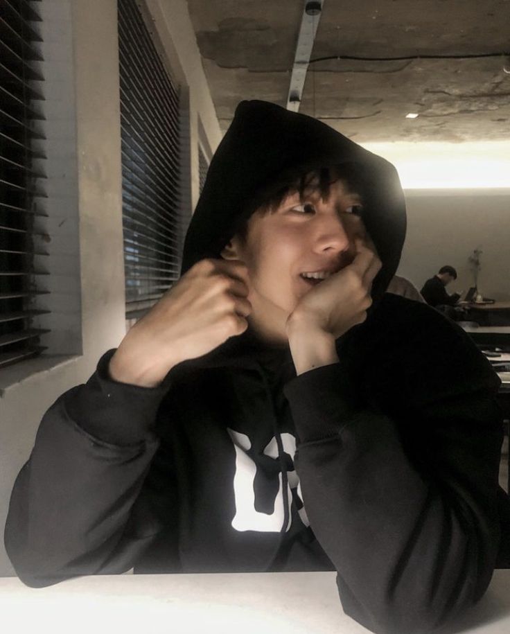 a young man sitting at a table with his hands on his face and wearing a hoodie