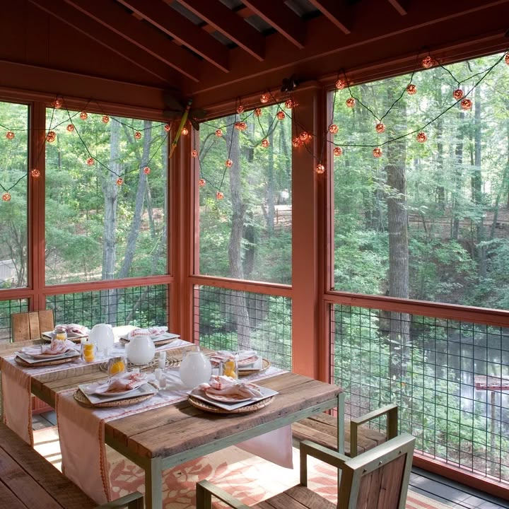 a dining room table set with place settings on the screened porch overlooking trees and woods