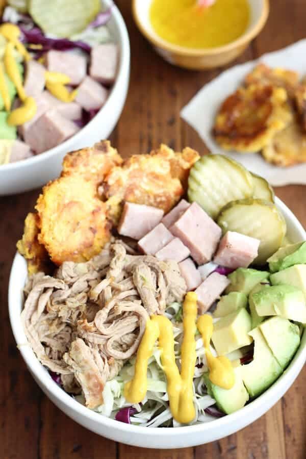 two bowls filled with different types of food on top of a wooden table next to bread and cucumber