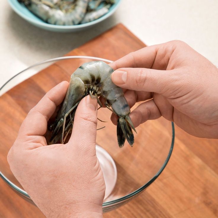 a person holding a shrimp in front of a glass bowl on top of a wooden table