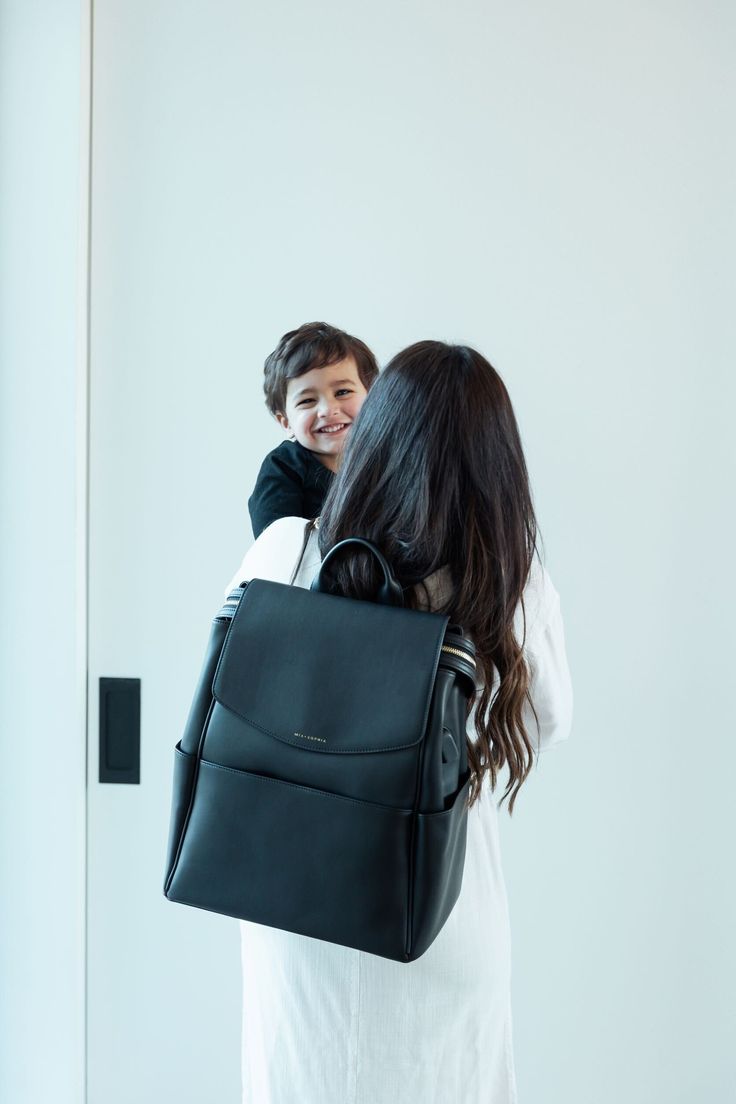 a woman holding a child in her arms and wearing a black backpack over her shoulder