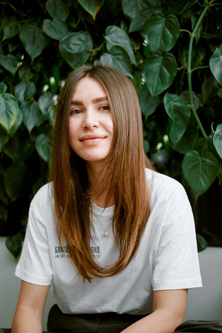 a woman with long brown hair sitting in front of a green plant and smiling at the camera