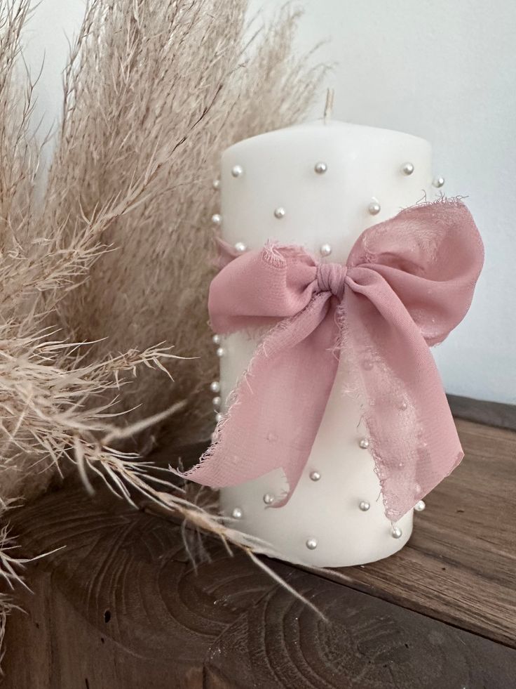 a white candle with a pink bow sitting on top of a wooden shelf next to dry grass