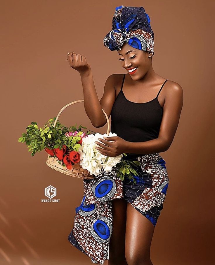 a woman holding a basket with flowers in it and wearing a black tank top, blue patterned skirt and head wrap