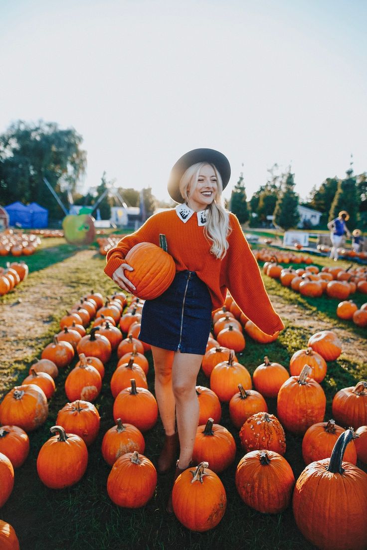 a woman in an orange sweater and black hat standing among pumpkins