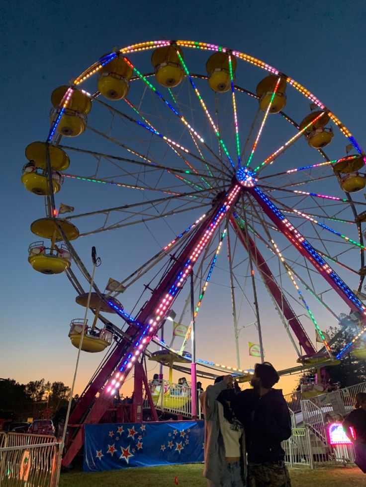 two people standing next to each other near a ferris wheel at night with lights on