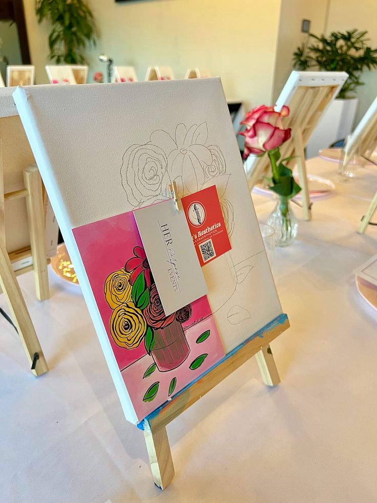 an easel with flowers and cards on it sitting on a white tablecloth covered table