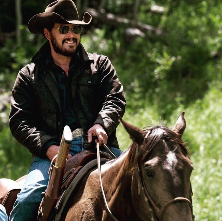 a man riding on the back of a brown horse next to a lush green forest