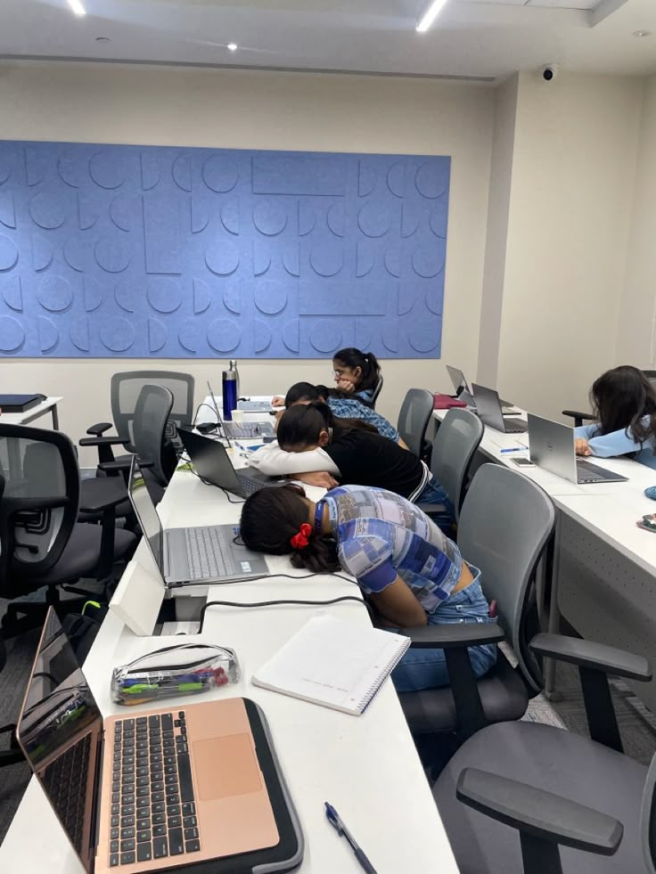 several people are sleeping on desks in an office setting with computers and laptops