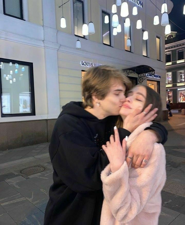 two people kissing each other in front of a building with lights hanging from the ceiling