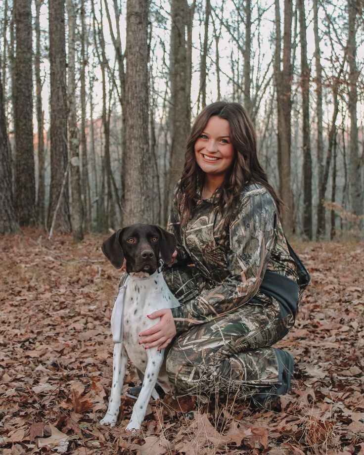 a woman kneeling down in the woods with her dog, who is wearing camo