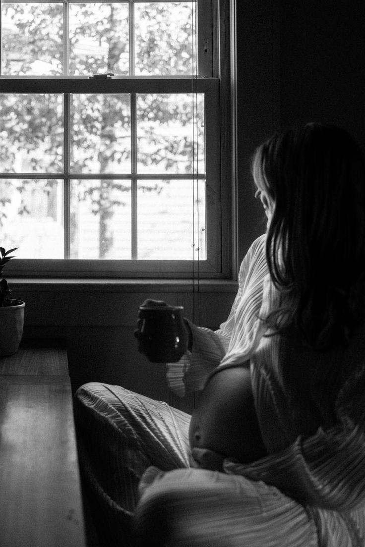 a pregnant woman sitting on a bed in front of a window holding a coffee cup