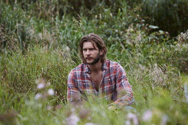 a man with long hair sitting in the middle of tall grass and looking off into the distance