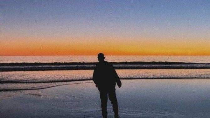 a man standing on top of a beach next to the ocean under a colorful sky