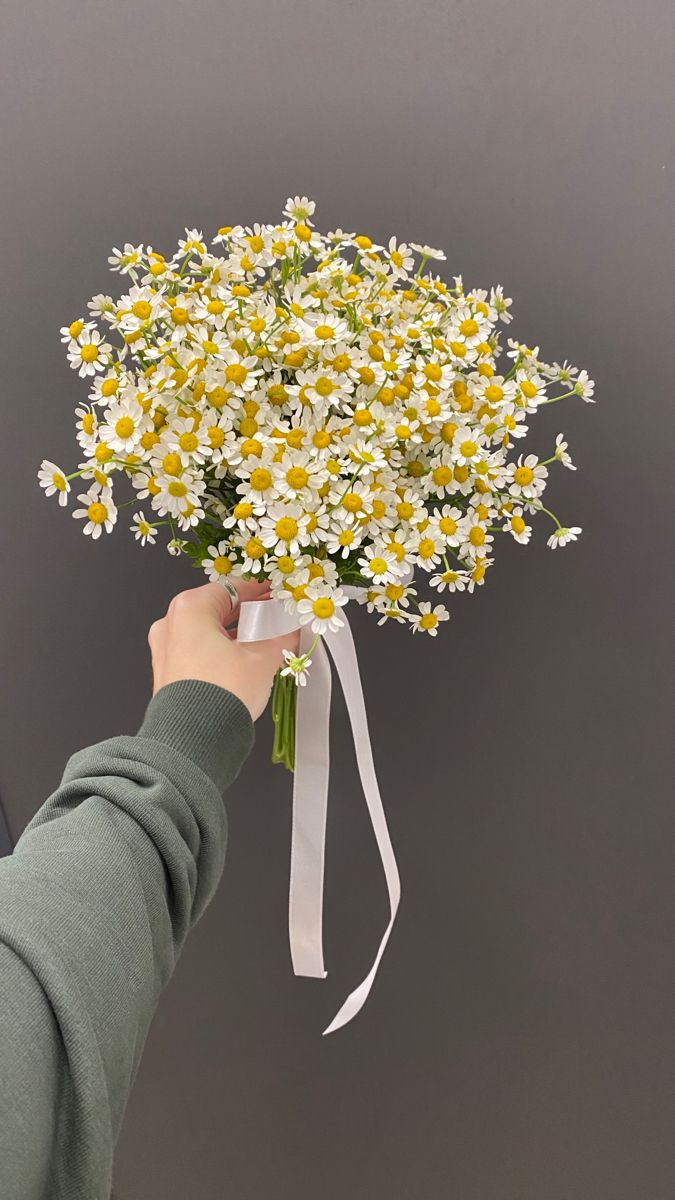 a person holding a bouquet of daisies in front of a gray wall with white ribbon