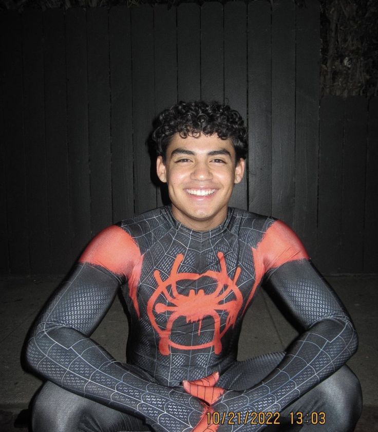 a young man in spiderman costume sitting on the ground with his hands crossed and smiling