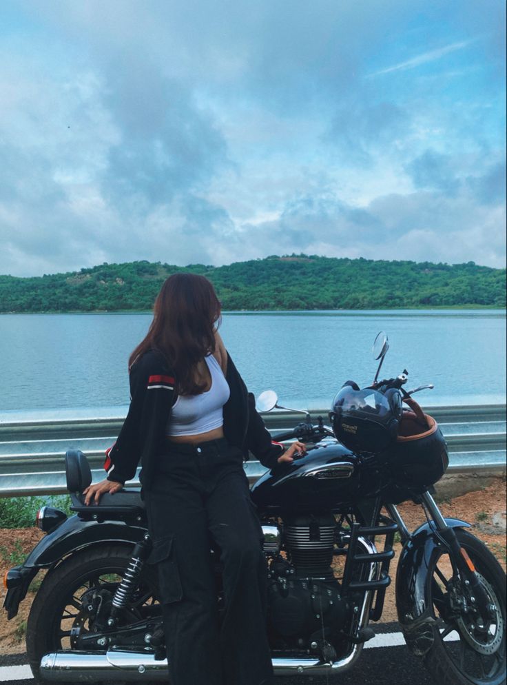 a woman standing next to a motorcycle on the side of a road near a body of water