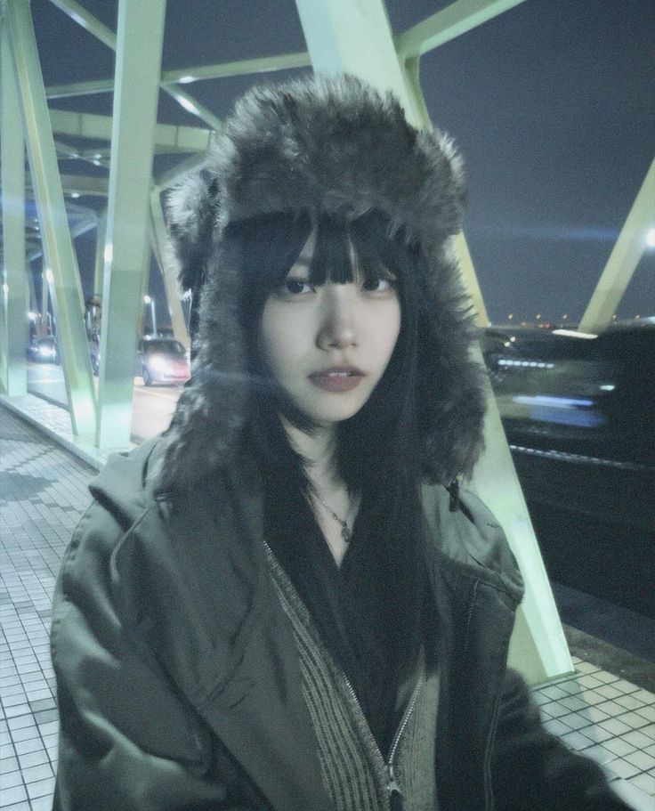 a woman wearing a fur hat and jacket standing on a bridge over looking at the camera