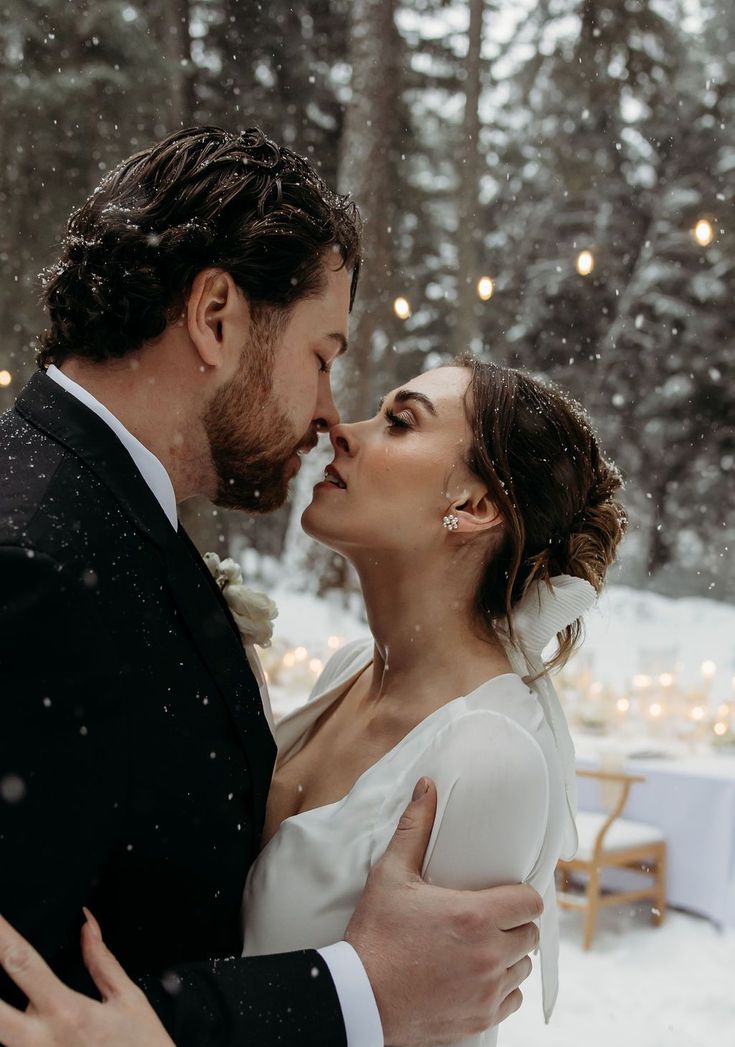 a bride and groom kissing in the snow at their winter wedding reception, surrounded by lights