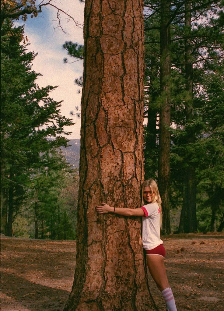 a woman standing next to a tall tree in the forest with her arm on it's trunk