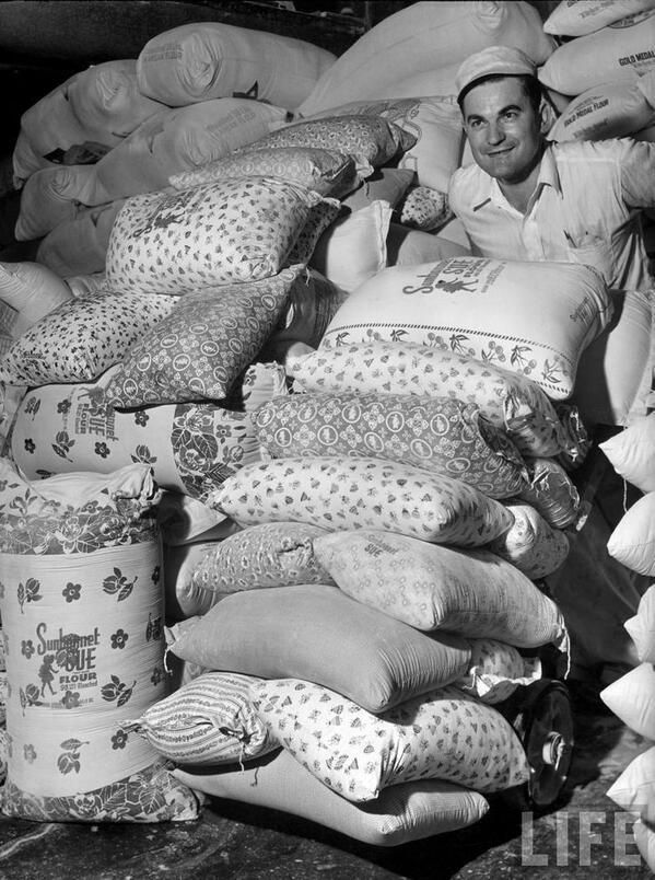 black and white photograph of a man in front of piles of bags with cats on them