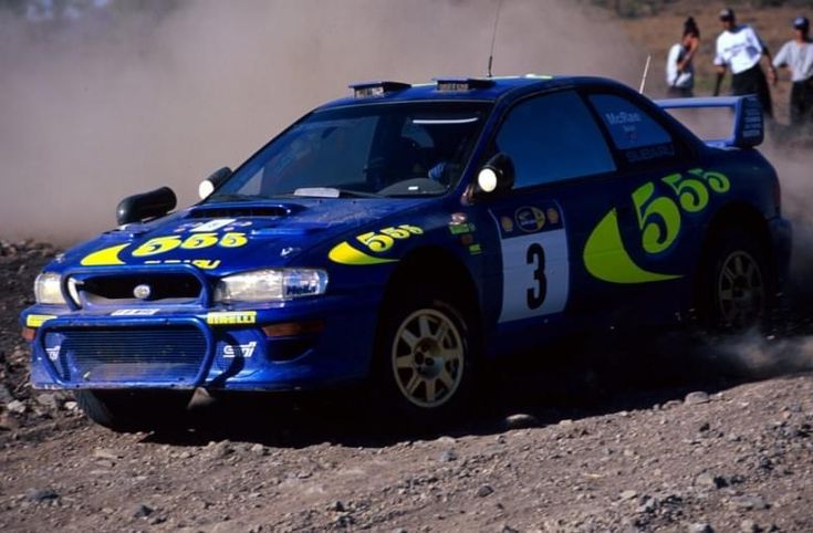 a blue car with yellow numbers on it driving down a dirt road in front of people