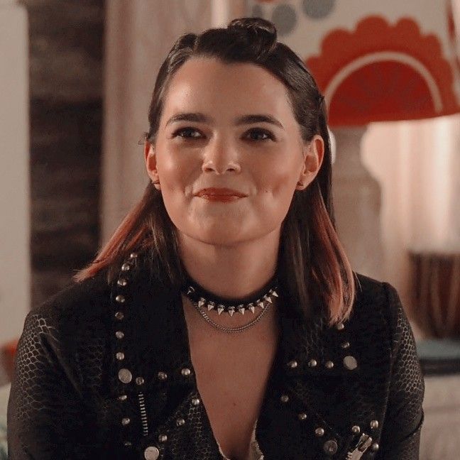 a woman with long hair wearing a black jacket and pearls on her neck is looking at the camera