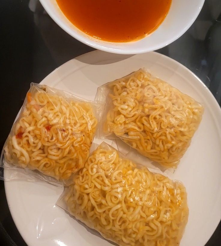 three bags of noodles on a white plate next to a bowl of soup and sauce