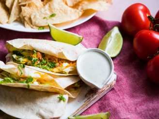 three tacos on plates with tomatoes, limes and sour cream in the background