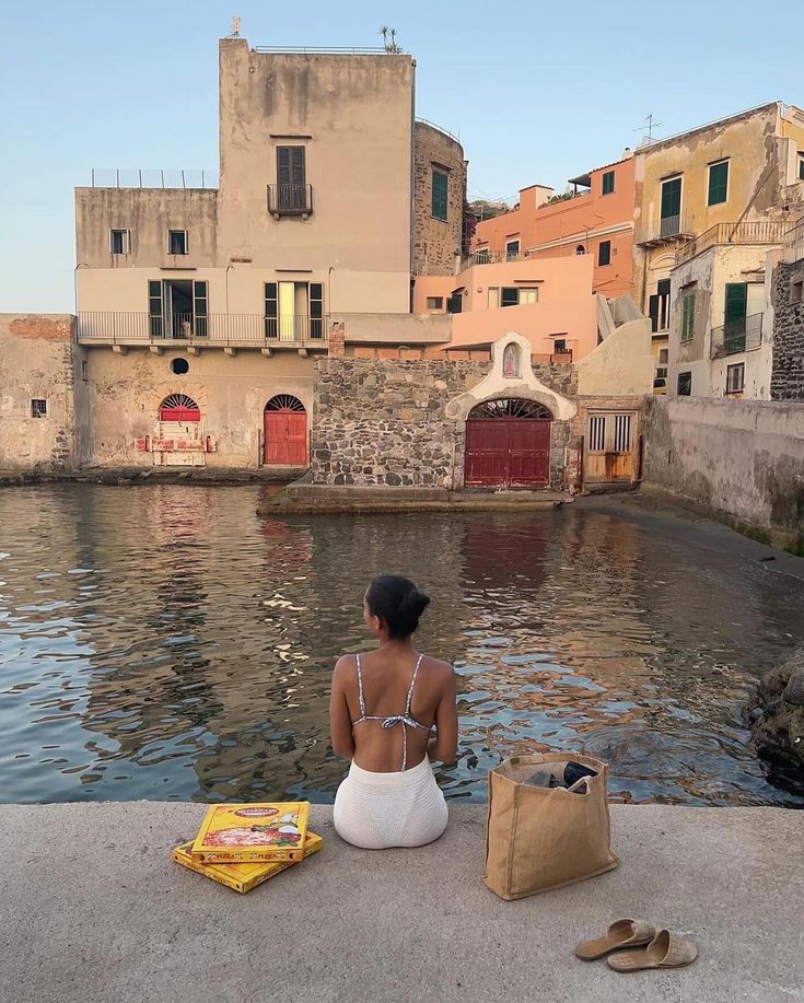 a woman sitting on the edge of a body of water next to buildings and a book