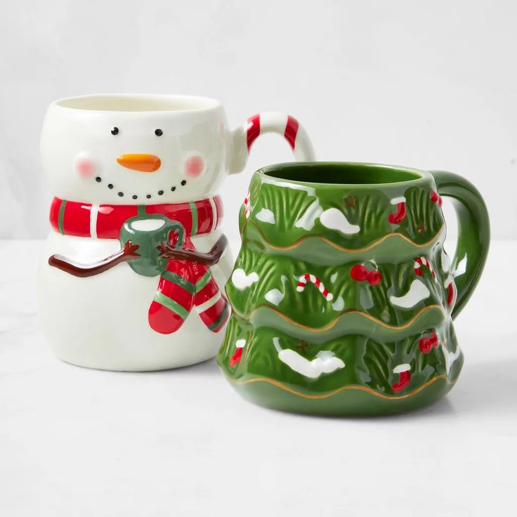 two ceramic mugs decorated with snowmen and christmas trees, one is green and the other is white